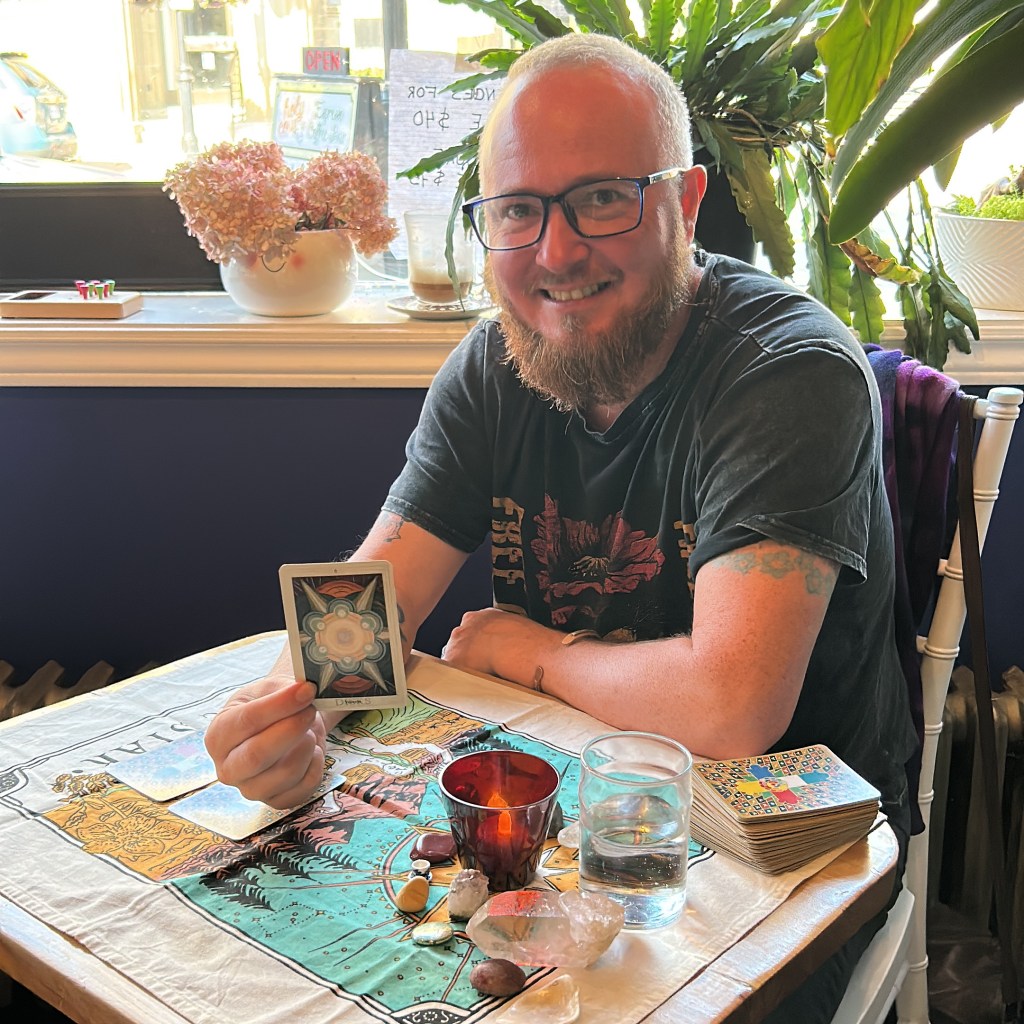 Hanlon sits in a window holding up the six of discs tarot card, he smiles at the camera. Hanlon beams because he knows the day is going well. He's a white man with a reddish beard, receding hairline and salt and pepper hair. The plant behind him gives the feeling of a green and lush moment in time.